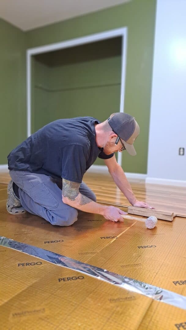A person wearing a cap and glasses is kneeling on a floor in front of an unfinished wall, installing wooden floor planks with various tools.